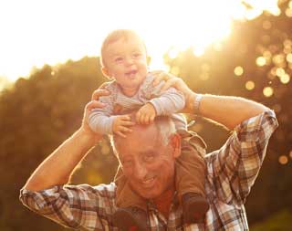 A smiling child being carried on his grandfather's shoulders in a park at sunset