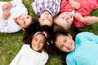 Five smiling children laying in the grass, their heads touching, looking up at the camera