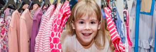 A young girl smiles as she peeks out from between several items of clothing hanging on a rack
