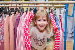 A young girl smiles as she peeks out from between several items of clothing hanging on a rack