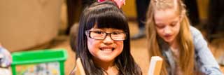 A young girl holding building blocks smiles at the camera while another girl plays behind her