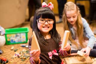 A young girl holding building blocks smiles at the camera while another girl plays behind her