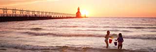Two young girls play in Lake Michigan while the sun sets behind them, outlining a lighthouse
