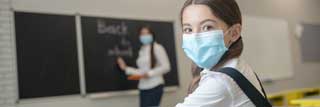 A young girl wearing a school uniform and protective mask sits in class while her teacher writes “Back to school” on the chalkboard.