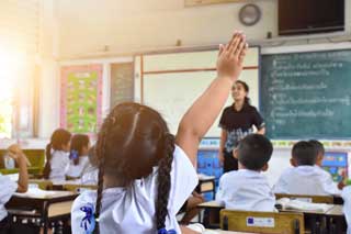 A young girl raises her hand to answer a question in a classroom full of students