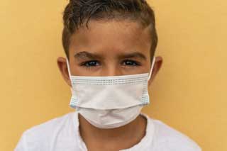 A young boy wearing a facemask standing in front of a yellow wall, looking at the camera