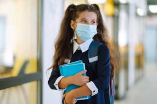 A student wearing a protective face mask and school uniform, carrying her books down a hallway