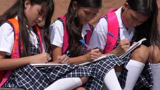 Three young girls in school uniforms sitting outside, taking notes