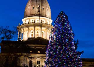 The exterior of the Michigan State Capitol building, with a Christmas tree in front of it, lit up at dusk.