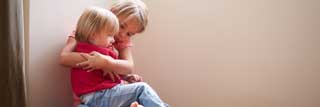 Two young children sitting in the corner, the older of the two comforting the younger who is sitting in her lap.