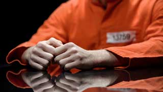 A prisoner in an orange jumpsuit with their hands resting on a highly-polished table.
