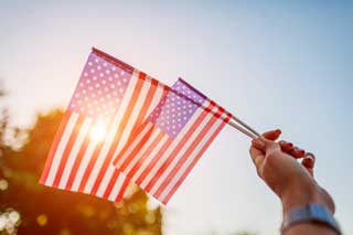 A woman's hand, holding two small American flags in front of a brilliant sunrise