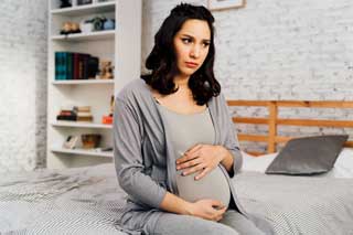 A concerned-looking pregnant woman sitting on her bed, protectively holding her belly
