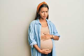 A concerned-looking pregnant woman leans against a wall while cradling her belly.