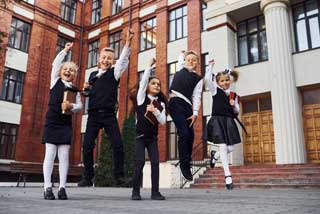 Five young children in school uniforms celebrating in front of their school.