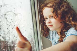 A young girl draws a heart in the raindrops that have collected on a window