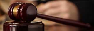 A judge's gavel resting on a table. The judge's hands are visible in the background, paging through a book.