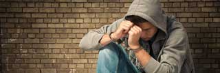 A youth in handcuffs holds his head in his hands while leaning against a brick wall