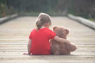 A little girl sits on a wooden walkway with her arm around a teddy bear