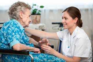 A young nurse smiles at an elderly woman in a wheelchair while handing her a glass of water
