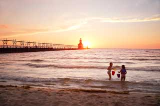 Two young girls play in Lake Michigan while the sun sets behind them, outlining a lighthouse