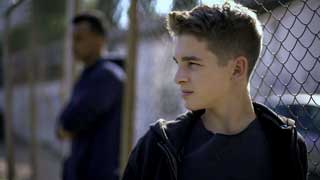 Two incarcerated young men lean against a chain link fence, staring into the distance