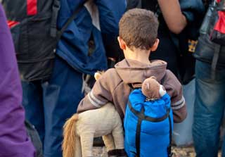 A young boy carrying a stuffed animal under one arm and another in his backpack waits in line