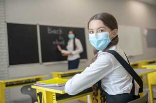 A young girl wearing a school uniform and protective mask sits in class while her teacher writes “Back to school” on the chalkboard.