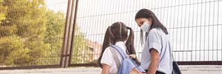 A young girl and her mother wearing protective face masks and walking hand in hand to school.