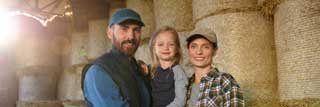 A husband, wife, and their daughter, standing in front of bales of hay and smiling