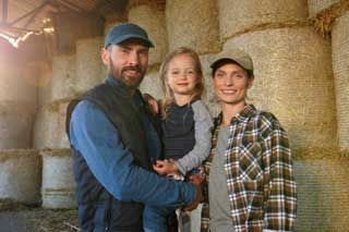 A husband, wife, and their daughter, standing in front of bales of hay and smiling