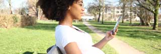 A young pregnant woman sitting on a park bench, reading on her phone.