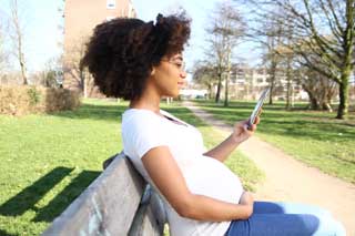 A young pregnant woman sitting on a park bench, reading on her phone.