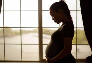A young pregnant woman outlined by the light coming in from the window behind her.