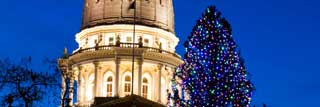 The exterior of the Michigan State Capitol building, with a Christmas tree in front of it, lit up at dusk.