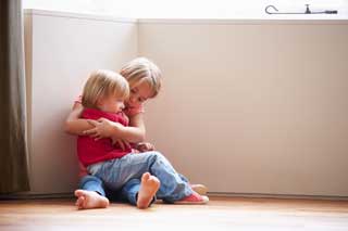 Two young children sitting in the corner, the older of the two comforting the younger who is sitting in her lap.