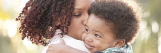 A mother holds her smiling child while kissing him on the cheek.