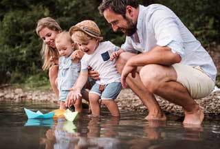 A happy family consisting of a mother, father, and two children standing at the edge of a river launching paper boats.