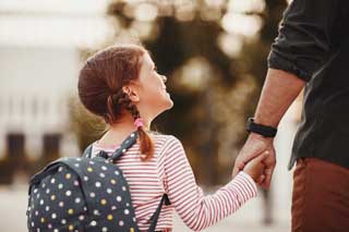 A young girl on the way to school, smiling and wearing a backpack, olding her father's hand.