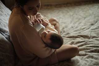 A mother sitting cross-legged on the bed, feeding her child from a bottle